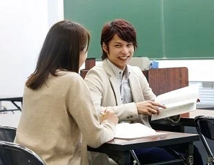 教室で男女二人が机を挟んで向かい合い、男性が本を開きながら笑顔で女性と話している様子