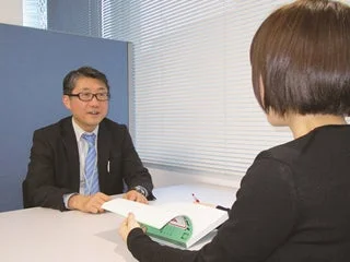 オフィスでスーツ姿の男性と女性がテーブルを挟んで向かい合い、書類を広げながら面談や打ち合わせをしている様子